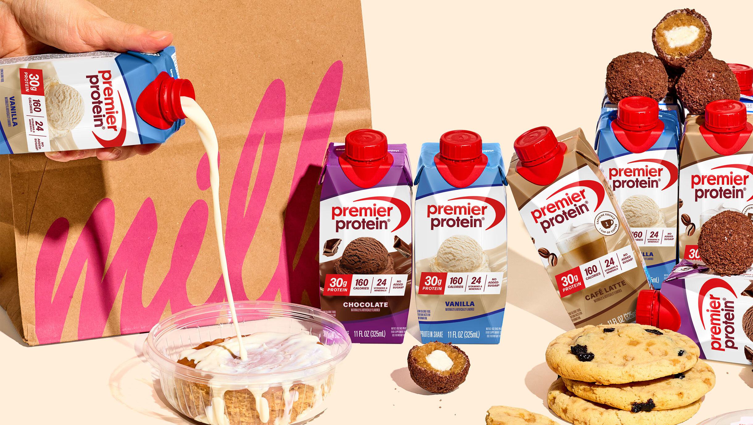 A collection of Milk Bar treats and Premier Protein shakes in front of a Milk Bar bag. A hand is pouring a Vanilla Protein Shake onto one of the treats.